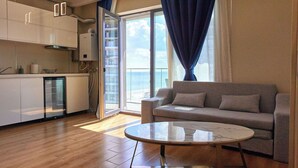 A modern living room with a sofa and a glass coffee table, a kitchen with white cabinets, and a large window with a view of the sea. - Signature Mamaia Nord (Mamaia-Sat)