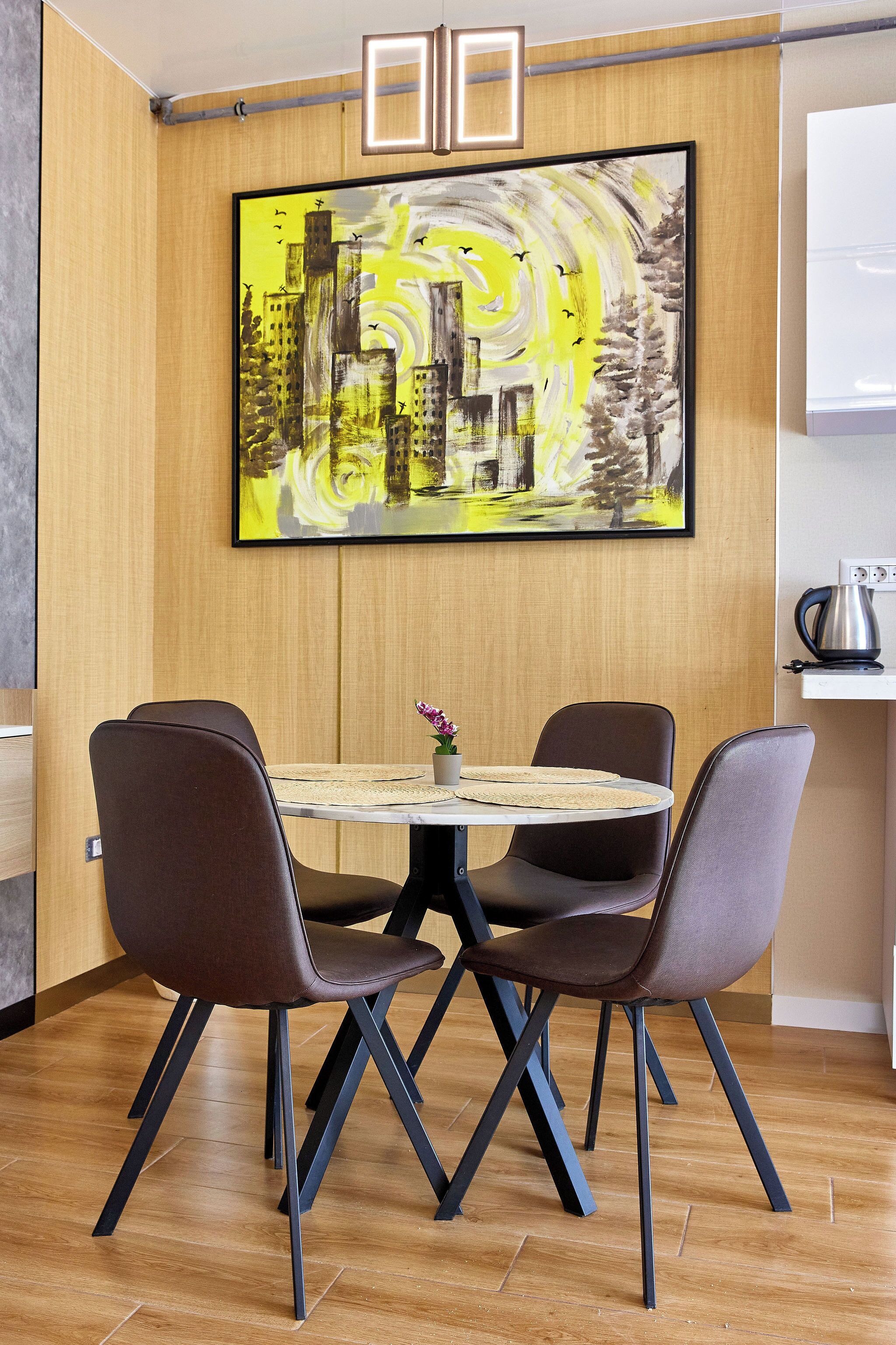 A dining area with a round table, four chairs, and a painting on the wall.