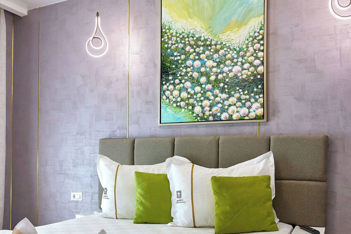 A modern hotel room with a large bed, a painting of flowers on the wall, and a unique light fixture above the bed.