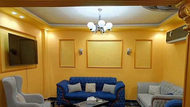 A living room with a blue sofa, a white armchair, and a grey sofa. There is a chandelier on the ceiling and a flat-screen TV mounted on the wall. - THE MESSAHA ROYALE (Giza Governorate)