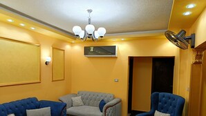 A living room with two blue sofas, a grey sofa, and a blue armchair. There is a chandelier, a wall-mounted air conditioner, and a floor fan. - THE MESSAHA ROYALE (Giza Governorate)