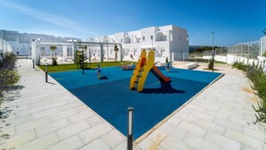 A playground with a slide and climbing structure, surrounded by white buildings and a clear blue sky.