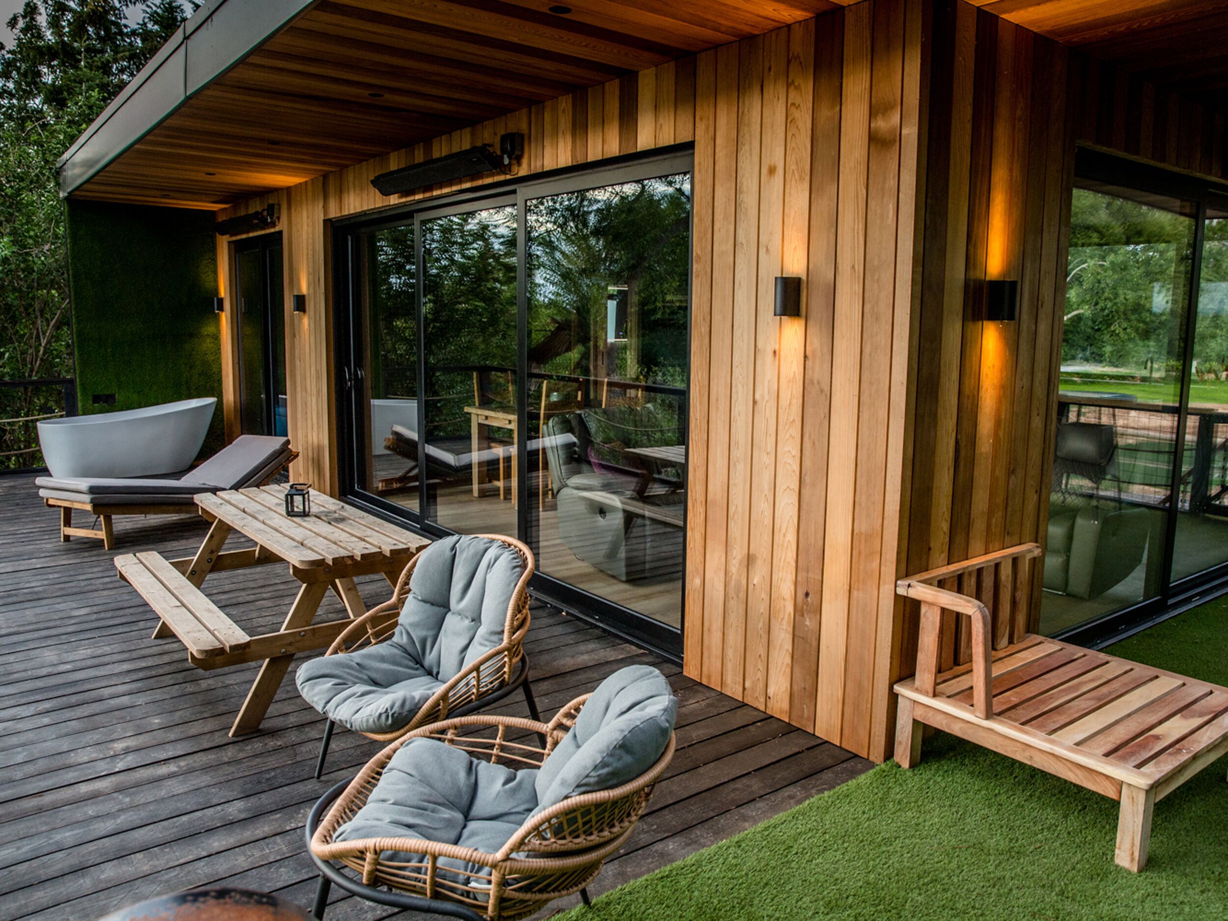 A wooden deck with wicker chairs and a lounge chair, a glass door leading to an outdoor area, and a wooden wall with a light fixture.