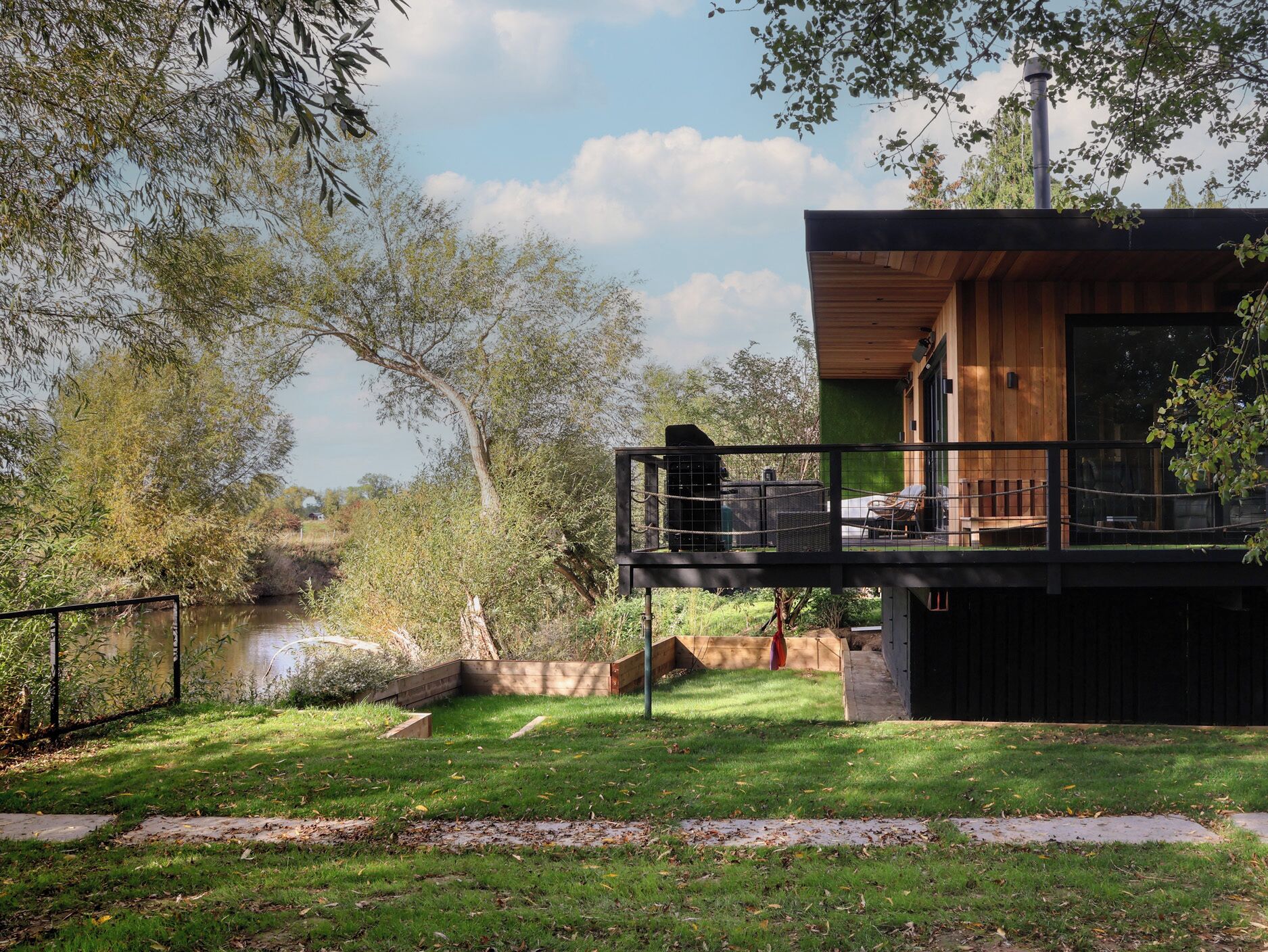 Une cabane moderne dotée d’une terrasse donnant sur une rivière, le tout entouré d’une végétation luxuriante.