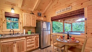Interior de una cabaña de madera con cocina, comedor y ventana con vistas a la vegetación.