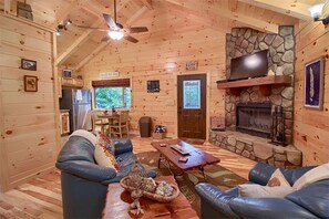 A cozy cabin interior with a stone fireplace, leather sofas, a dining area, and a ceiling fan.