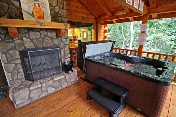 A hot tub with a stone fireplace, wooden deck, and a view of the surrounding forest.