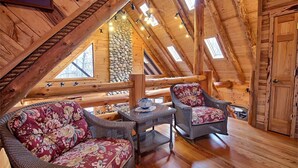 Ein gemütliches Dachzimmer mit Holzbalken, einer Steinwand und zwei Korbstühlen mit Blumenkissen.