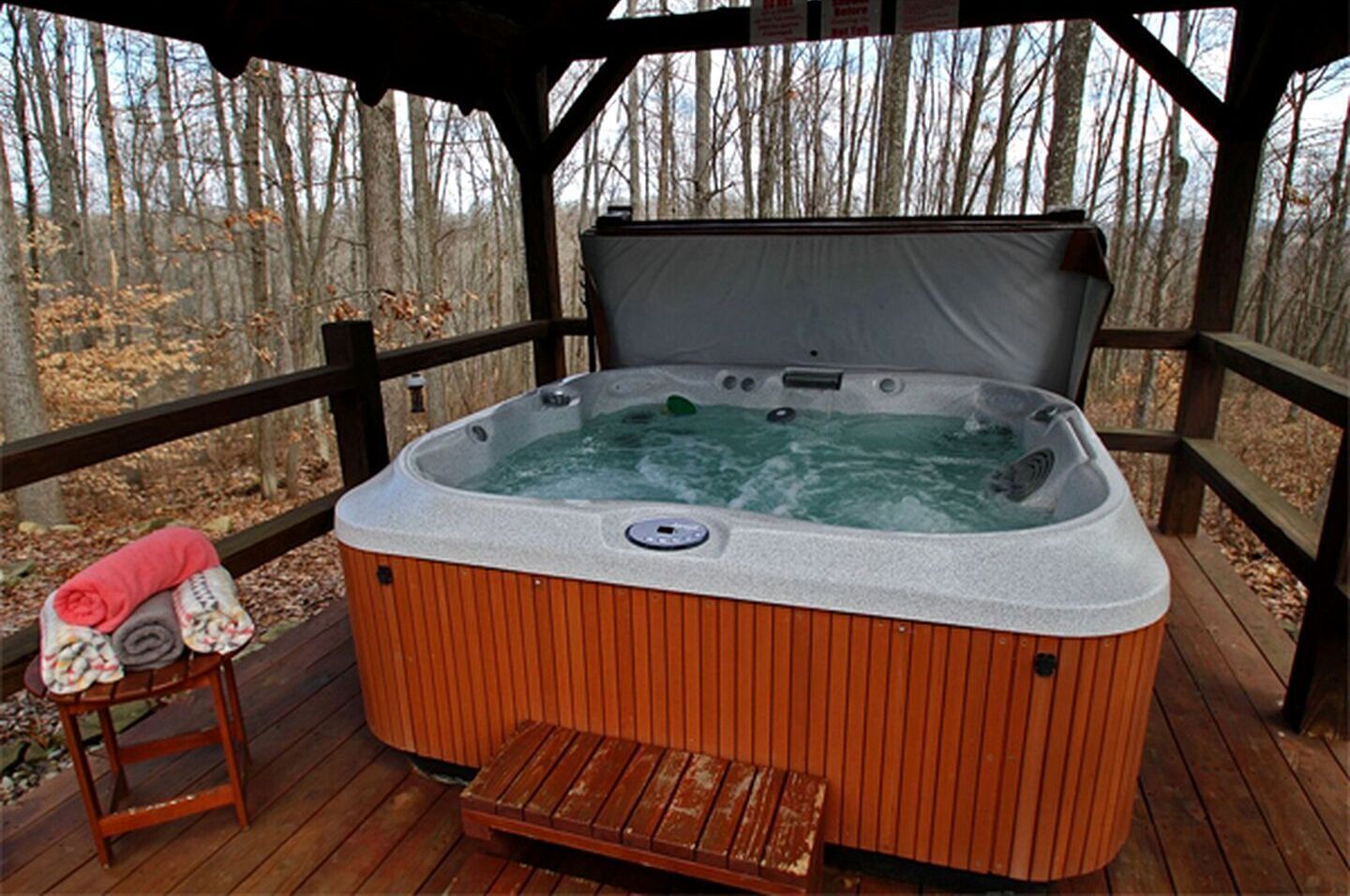 Un jacuzzi en una terraza de madera con un banco y toallas, rodeado de árboles.