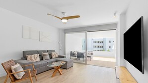 A modern living room with a grey sofa, a glass coffee table, and a woven chair. There is a ceiling fan, a large TV, and a sliding glass door leading to an outdoor balcony. - Ref. 585392 (Motril)