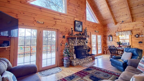 A log cabin interior with a stone fireplace, wooden walls, and large windows.