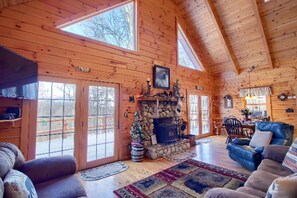 A log cabin interior with a stone fireplace, wooden walls, and large windows.