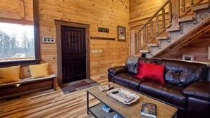 L’intérieur chaleureux d’un chalet en bois comprend un canapé en cuir, un plancher en bois, un escalier et une fenêtre donnant sur un paysage enneigé.
