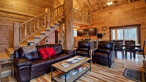 L’intérieur d’une cabane en rondins, avec un escalier en bois, des meubles en cuir, un coin cuisine et de grandes fenêtres.