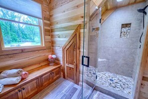 A wooden cabin interior with a glass-enclosed shower, a window with a view of trees, and a wooden bench with towels.