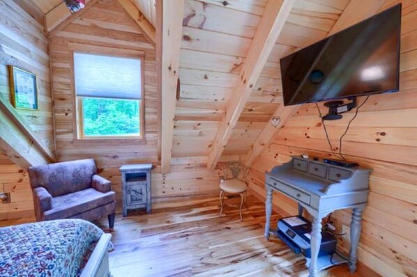 A log cabin room with a bed, a chair, a small table, a TV, and a window with a view of greenery.