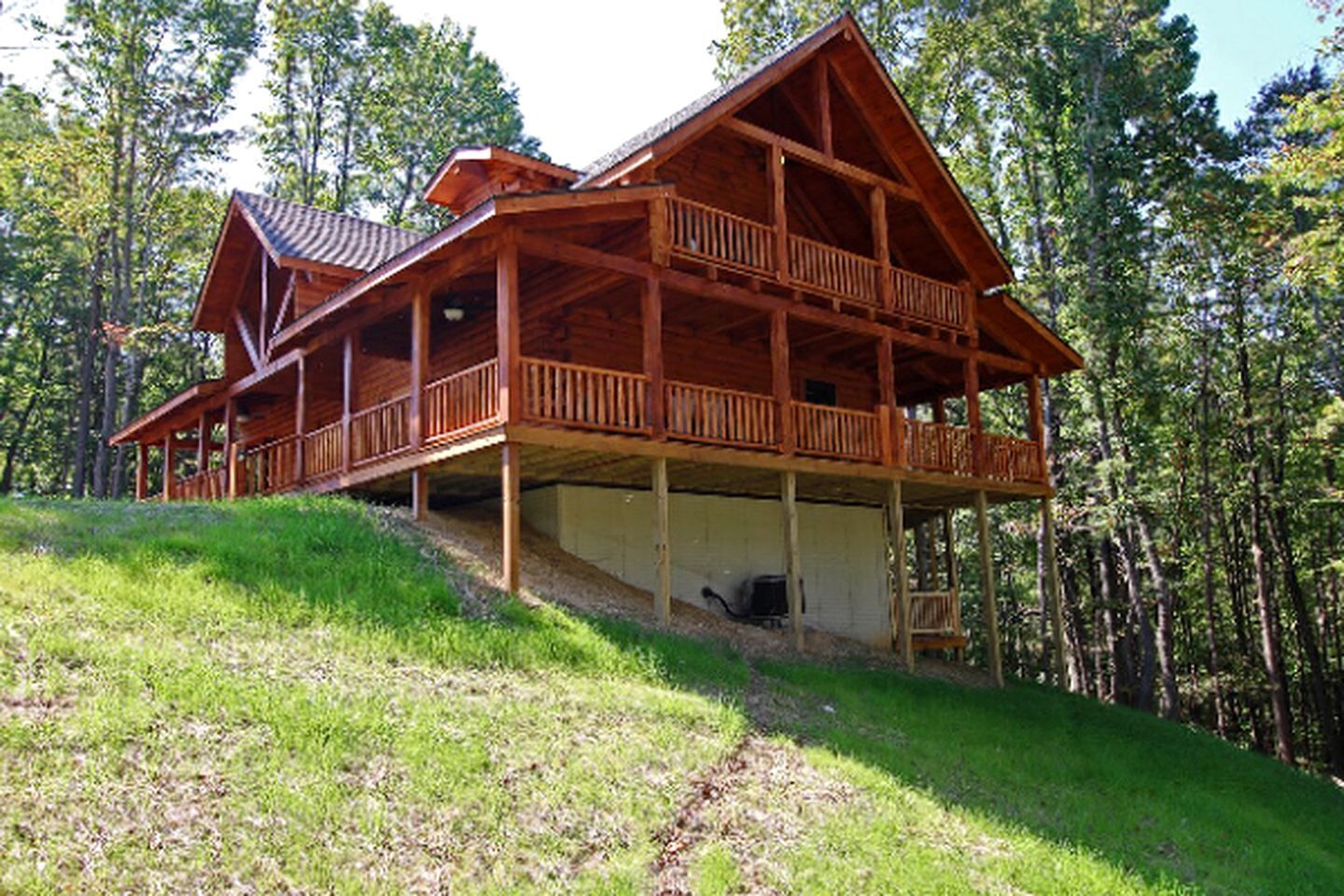 A wooden cabin with a balcony, set on a grassy hill surrounded by trees.