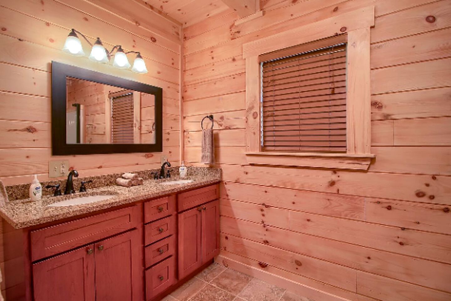Une salle de bain rustique, avec des murs en bois, un lavabo en granit, un miroir avec éclairage intégré et une fenêtre équipée de stores.