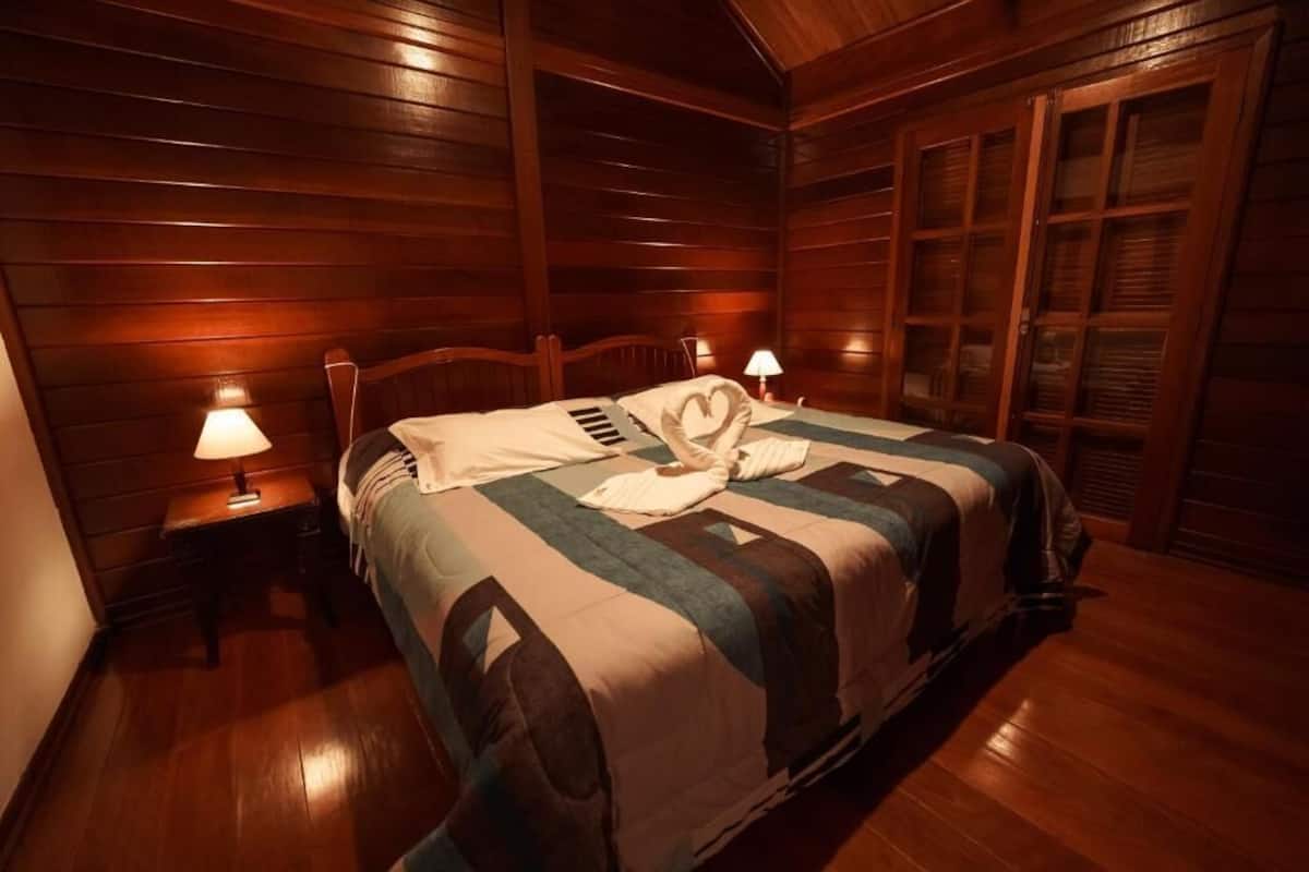 A wooden-paneled room with a bed, two bedside lamps, and a window with blinds.