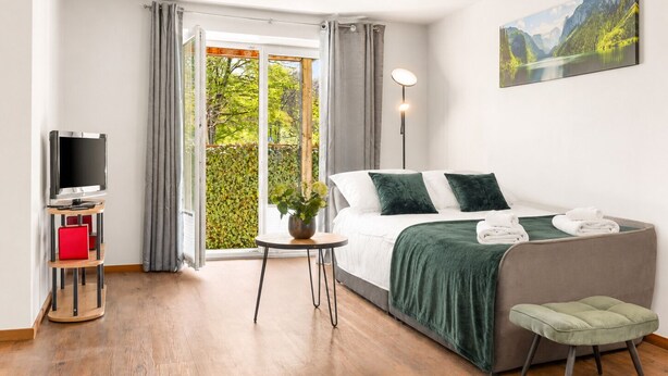 A modern hotel room with a bed, a TV, a small table with a plant, and a painting on the wall.