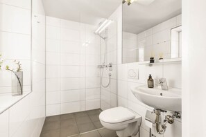 A modern bathroom with white tiles, a glass shower enclosure, a toilet, a sink with a mirror, and a small shelf with toiletries.