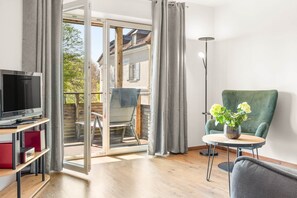 A room with a television, a table with a vase of flowers, a green chair, and a sliding glass door leading to an outdoor area.
