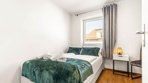 Un dormitorio con cama, una mesita de noche, una lámpara, una ventana con cortinas y vista a un edificio.