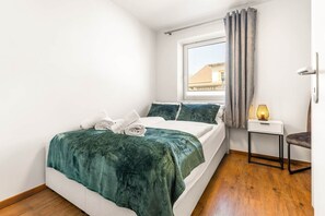 Un dormitorio con cama, una mesita de noche, una lámpara, una ventana con cortinas y vista a un edificio.