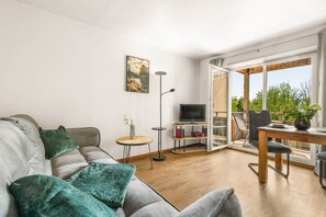 Una sala moderna con un sofá gris, una mesita auxiliar pequeña, una lámpara de pie, un televisor sobre un mueble bajo y un balcón con una mesa y sillas de madera.