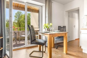 A modern kitchen with a wooden table set for two, a dining chair, and a view through the glass door to an outdoor area with a wooden deck and chairs.
