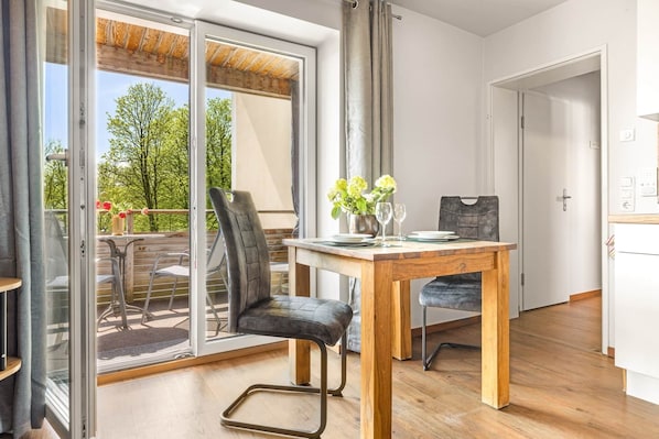 A modern kitchen with a wooden table set for two, a dining chair, and a view through the glass door to an outdoor area with a wooden deck and chairs.