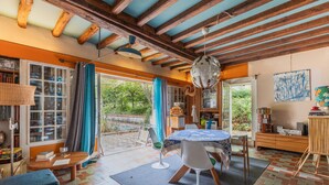 A room with a wooden ceiling, a round table with chairs, a bookshelf, and a view of greenery outside.