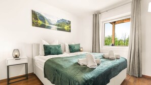 Un dormitorio con cama, mesita de noche, lámpara, ventana con vistas y un cuadro en la pared.