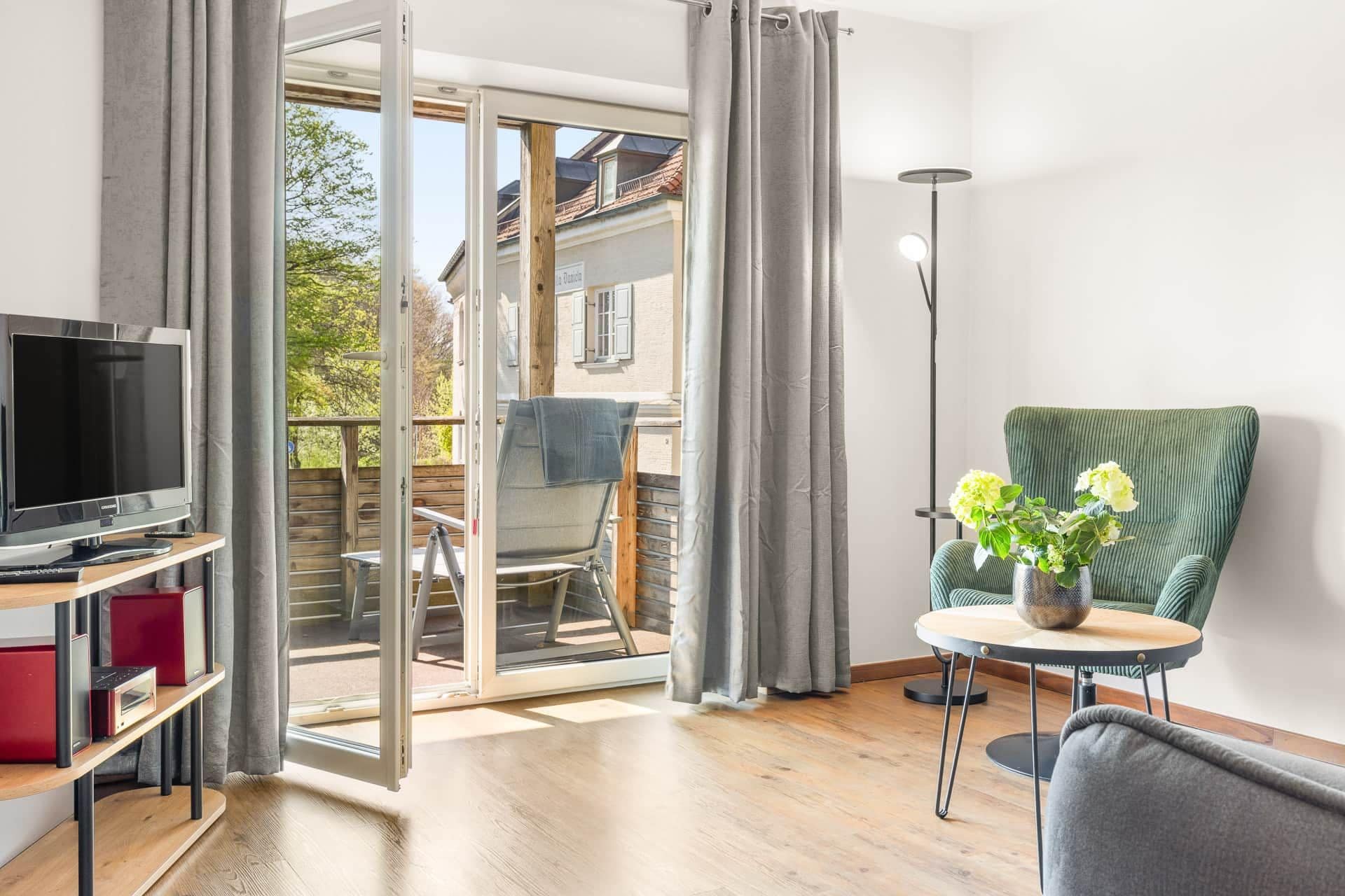 A room with a television, a table with a vase of flowers, a green chair, and a sliding glass door leading to an outdoor area.