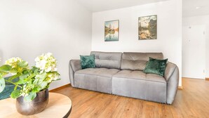 Un sofá gris con dos cojines verdes, una mesa redonda de madera con una planta en maceta y dos cuadros en la pared.
