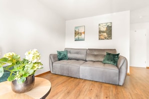 A grey sofa with two green cushions, a round wooden table with a potted plant, and two framed pictures on the wall.