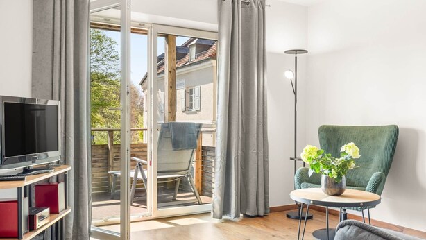 Una habitación con televisión, una mesa con un florero, una silla verde y un ventanal corredero que da a un espacio exterior.