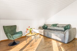 A loft-style room with a green upholstered chair, a grey sofa, and a small round table with a vase of flowers.