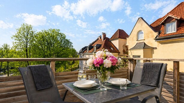 An outdoor dining area with a table set for two, a vase of flowers, and a view of buildings in the background.