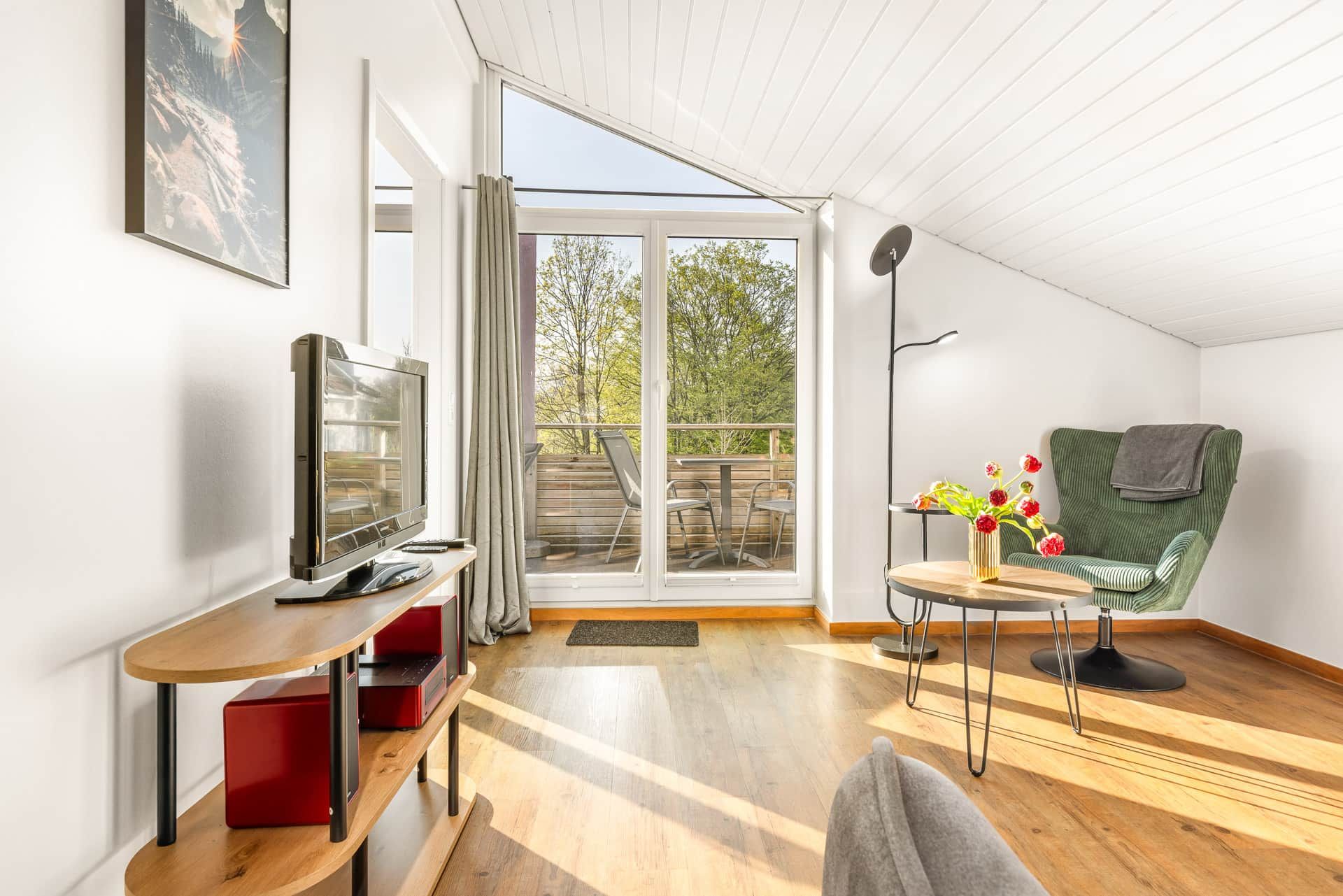 Uma sala de estar moderna com televisão, sofá, uma mesa pequena com flores e uma varanda com mobiliário exterior.