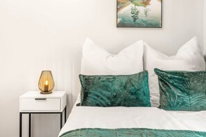 Un lit bien fait, agrémenté d’oreillers blancs et turquoise, une table de chevet blanche avec une lampe, et un tableau accroché au mur.