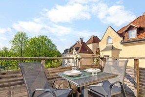 An outdoor dining area with a table set for two, surrounded by wooden railings, overlooking a residential neighborhood with houses and trees.