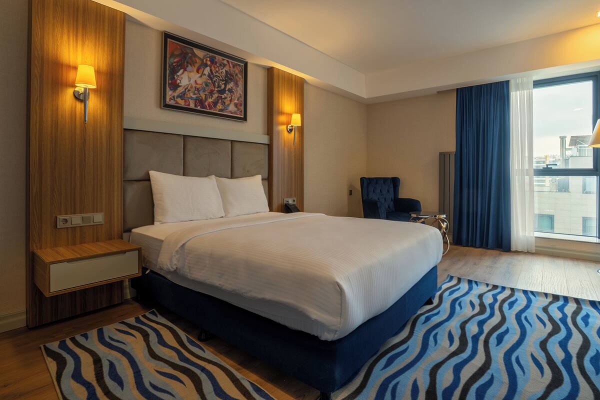 Habitación de hotel con una cama grande, una silla azul, una mesita de noche de madera, un cuadro en la pared y vista a la ciudad por la ventana.