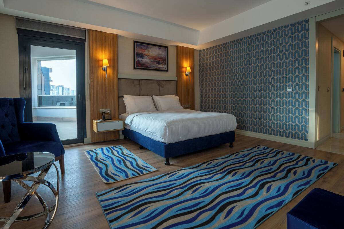 Una habitación de hotel moderna con una cama grande, un área de descanso con una silla, una mesita auxiliar y una alfombra estampada.