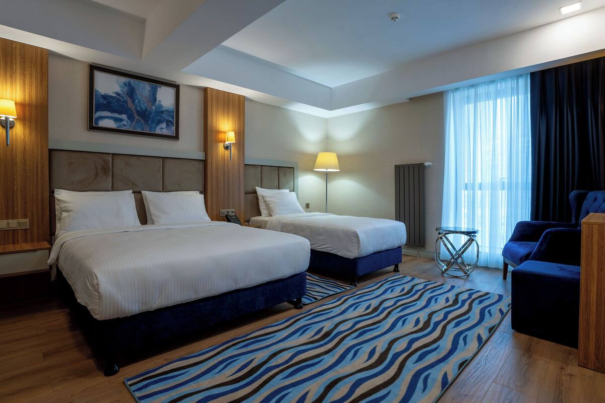 Habitación de hotel con dos camas, un sofá azul, un tapete estampado de cebra y un ventanal con cortinas.