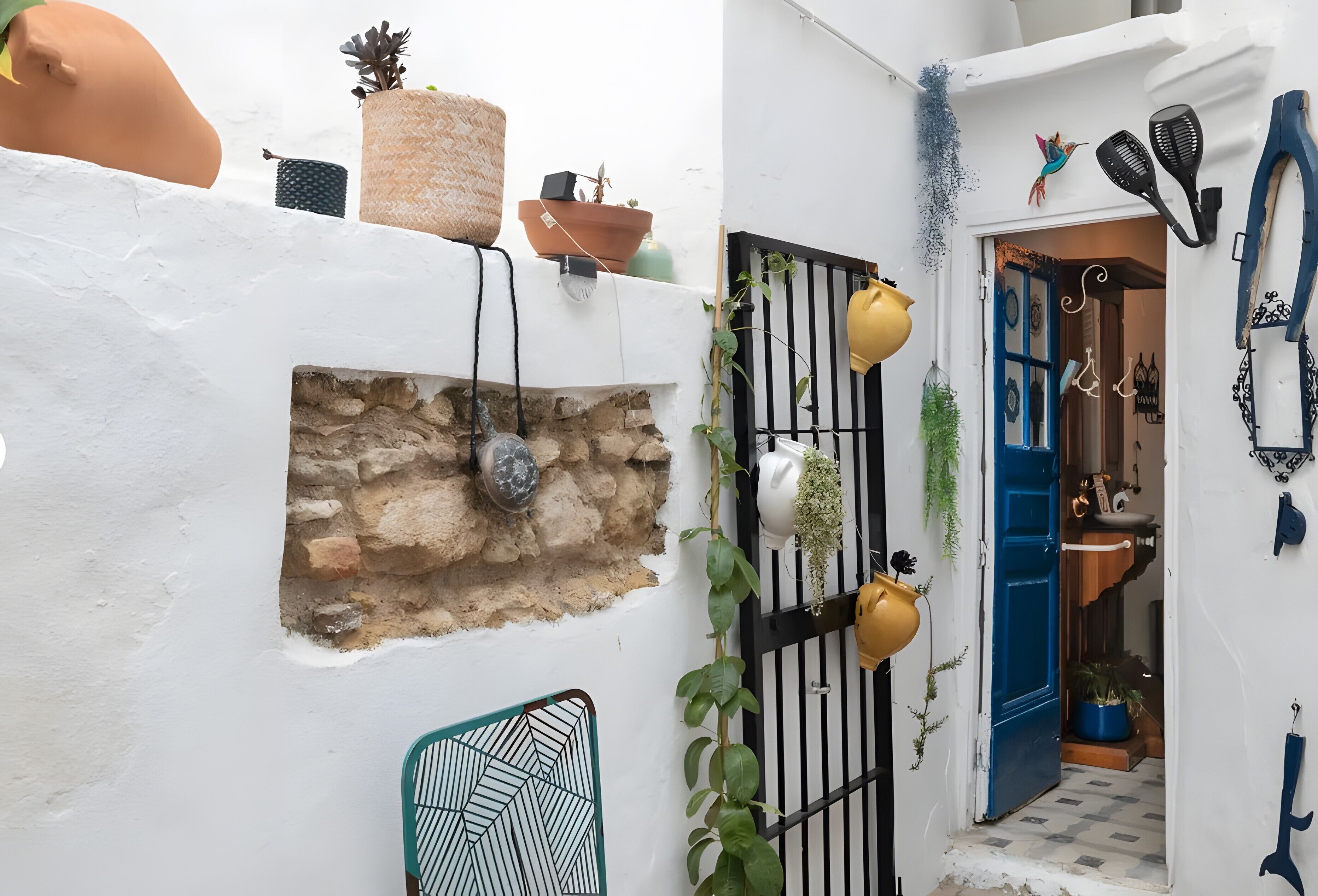 A narrow alleyway with a white wall, a stone wall, and a blue door. There are potted plants, a yellow pot, and a metal chair.