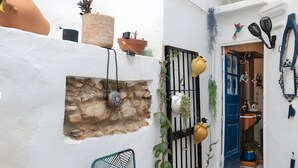 Une ruelle étroite avec un mur blanc, un mur de pierre et une porte bleue. On y trouve des plantes en pot, un pot jaune et une chaise en métal.