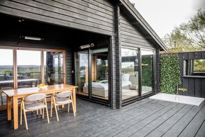 Un espacio moderno para comer al aire libre con una mesa y sillas de madera, un dormitorio con paredes de vidrio y vistas del paisaje circundante.