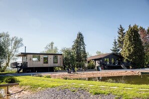 Une cabane moderne dotée d’une terrasse et d’un espace abrité, entourée d’un étang et d’arbres.