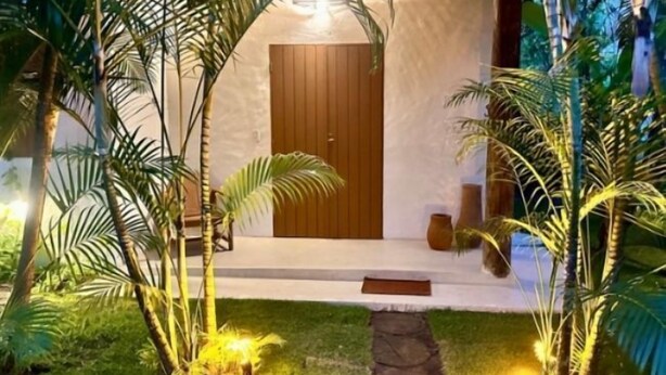 A well-lit entrance with a wooden door, surrounded by lush greenery and a stone pathway.